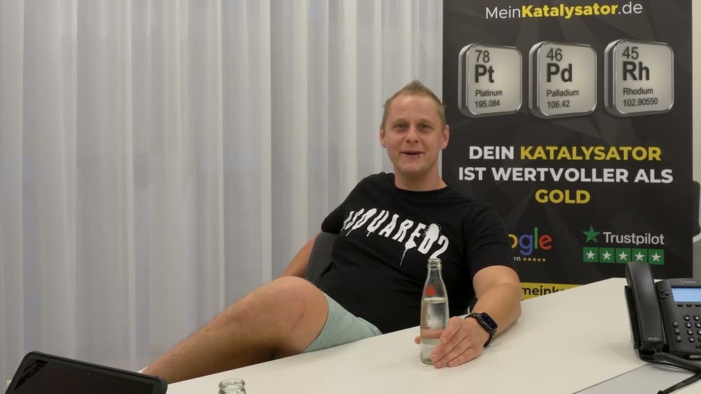 Mann in schwarzem T-Shirt sitzt am Schreibtisch mit Wasserflasche; Plakat "DEIN KATALYSATOR IST WERTVOLLER ALS GOLD"
